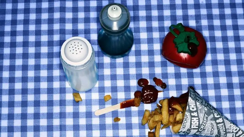 paper cone of potato chips pictured next to ketchup bottle, salt and pepper shakers on a blue checkered tablecloth