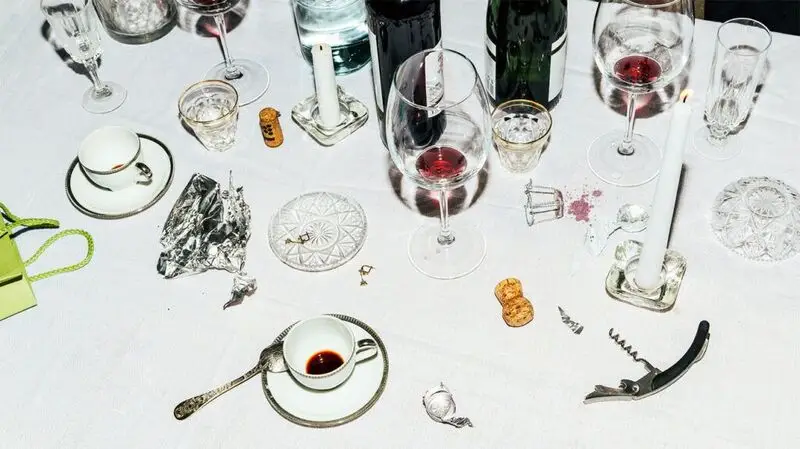 Empty shot and wine glasses and tea cups on a table, with red wine stains ona white table cloth, and candles