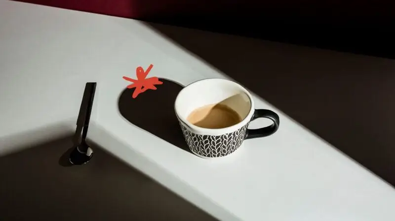 A cup of coffee and a spoon on the side sits on a flat surface, its shadow extending behind it