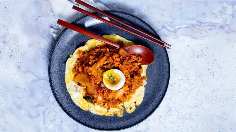 A plate of kimchi rice topped with an egg, with chopsticks and a spoon on the side of the plate