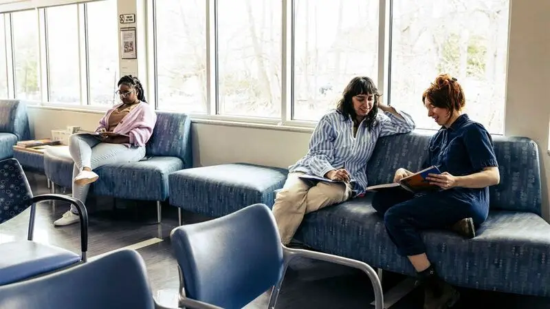 Two female patients fill out medical forms while one of them talks to a nurse on a sofa