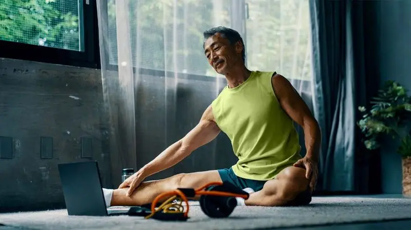 older Asian man stretching on the floor in front of a laptop