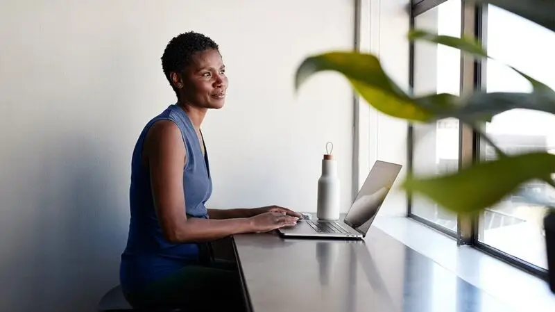 A woman looks out the window while sitting and typing on her laptop at a windowside table
