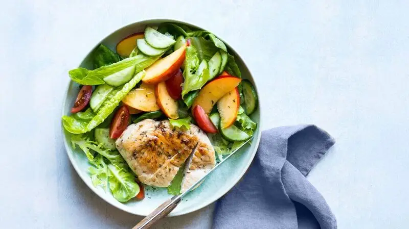 A bowl of grilled chicken salad with greens, peaches, cucumbers, and tomatoes