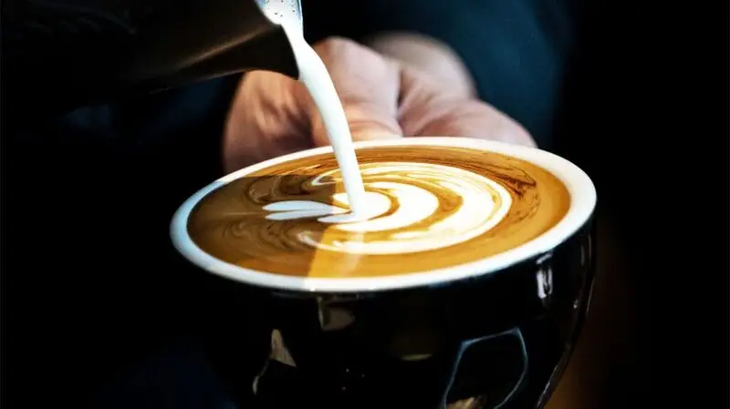 A person pours milk into and creates latte art in a cup of coffee