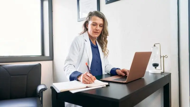 A doctor taking notes while working on a laptop.