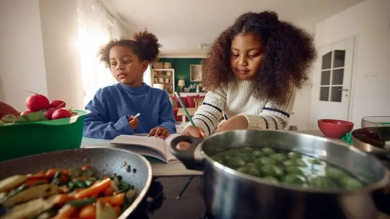 Two children do homework at a kitchen counter as someone prepared them a healthy meal with various vegetables