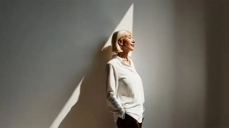 An older woman stands with her body slightly angled sideways against a wall as a ray of light illuminates her face
