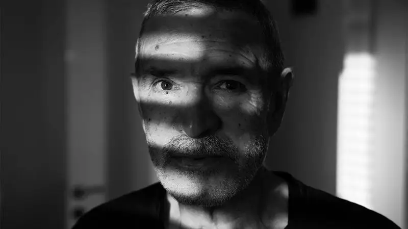 An older adult looking straight ahead as light from window blinds casts shadows on their face.