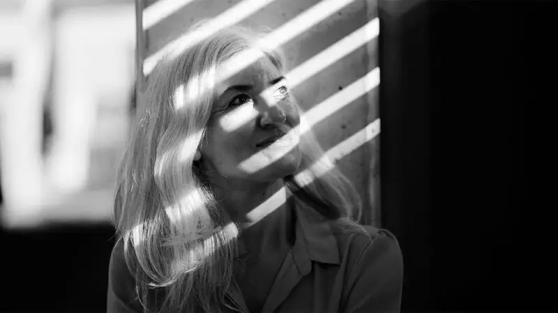 An older adult woman looks diagonally up as the light from the window blinds casts shadows on her face
