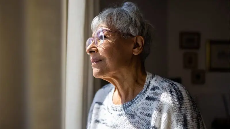 An older adult woman looks out of the window pensively sunlight shining on half her face
