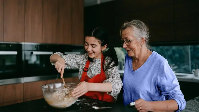 A young girl cooks with her grandmother in the kitchen, stirring food in a glass bowl
