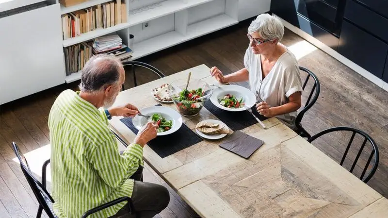 Two older adults having a meal of salad, crackers, cheese, and almonds for a gut microbiome-diverse diet at a table inside a home