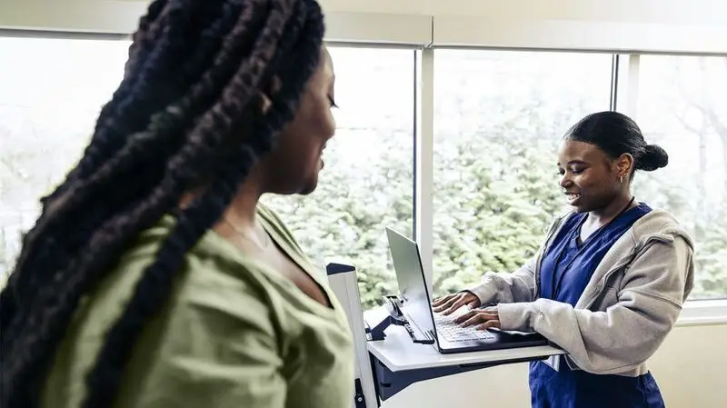 An oncology nurse takes notes on a laptop as a patient prepares for a mammogram to screen for breast cancer