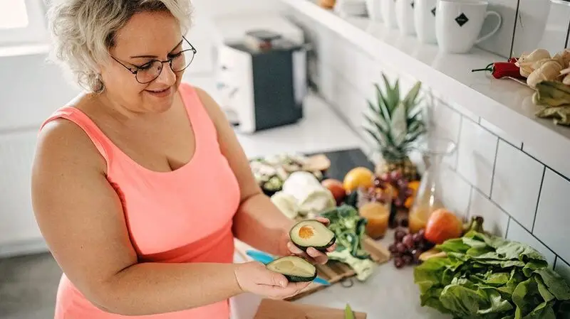 middle-aged white woman holding a sliced avocado in her kitchen
